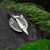 Back view of a unique silver Spartan shield pendant with sword detail, displayed on natural dark stone with lush greenery.