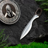 Close-up of handcrafted silver Spartan shield pendant and separate sword on dark stone, with green moss for natural contrast.